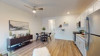 This single-floor unit features a thoughtfully designed open-concept layout that seamlessly connects the living, dining, and kitchen areas. Hardwood-style flooring runs consistently throughout, lending warmth and visual continuity to the space. The living area is anchored by a large window that invites natural light and is equipped with a ceiling fan for added comfort.

The kitchen is well-appointed with white shaker-style upper cabinets, gray lower cabinets, granite-look countertops, and a full suite of stainless steel appliances, including a range, over-the-range microwave, and refrigerator. Open wall shelving adjacent to the kitchen provides practical display and storage space.

The dining area accommodates a round table with seating for multiple guests and flows naturally from both the kitchen and living room, making the layout ideal for everyday use and entertaining.

The bedroom is generously sized, featuring multiple windows that frame lush outdoor greenery, a ceiling fan, and a distinctive sliding barn door that adds a modern-industrial touch. The room offers ample space for a full furniture arrangement.

The bathroom includes a granite-topped vanity with storage cabinetry, a shower/tub combination, and wood-look tile flooring, maintaining the consistent aesthetic found throughout the unit.