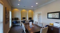 This single-floor space combines practicality and refined design in a well-appointed layout. The standout feature is a business center area where three arched wall niches, illuminated with warm yellow accent lighting, house individual computer workstations. Each alcove is finished with a built-in desk surface and complemented by stylish patterned chairs, creating a focused and comfortable work environment. The arched architectural detailing adds a classic, elegant touch to the room's overall aesthetic.

Adjacent to the workstations is a dedicated conference or meeting area, centered around a large, dark-wood rectangular table surrounded by upholstered chairs in neutral tones. This arrangement provides ample seating for group meetings or collaborative work sessions. A classic French door with grid-pane glass panels allows natural light to filter into the space while offering a visual connection to the exterior.

Recessed lighting in the white ceiling ensures bright, even illumination throughout, while crown molding along the ceiling line adds a polished, finished look. The neutral wall tones and patterned low-pile carpeting contribute to a professional yet inviting atmosphere, making this space well-suited for productivity and community engagement.