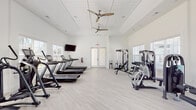 Located at Collins Road in Jacksonville, Florida, this single-floor fitness center offers a spacious and well-appointed environment designed to support a comprehensive workout experience. The interior is finished with a clean, modern aesthetic throughout, characterized by crisp white walls, light wood-look flooring, and soaring ceilings that lend an open, airy feel to the space.

The facility is thoughtfully divided into distinct workout zones. The cardio area lines one side of the room with multiple treadmills and elliptical machines, each equipped with individual monitors. A wall-mounted flat-screen television provides additional entertainment for cardio sessions. Dual ceiling fans keep the space comfortable, complementing the recessed lighting overhead.

The strength training section features a variety of cable machines, weight benches, a dumbbell rack, and selectorized resistance equipment, offering options for both isolated and compound exercises. Additional equipment includes a recumbent bike and stability tools such as balance balls and medicine balls.

Large windows along multiple walls flood the interior with natural light and provide views of the surrounding greenery, creating an inviting atmosphere. French doors offer convenient access. Practical touches like a fire extinguisher and emergency exit signage are also present throughout.