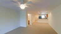 This two-floor space features an open, airy layout with wall-to-wall carpeting throughout the main living area. The living space is generously sized, offering a comfortable sense of openness accentuated by a ceiling fan with a light fixture overhead and recessed lighting that illuminates the area evenly. Neutral-toned walls and light-colored carpet contribute to a clean, bright aesthetic throughout.

A hallway leads toward additional rooms, with a doorway opening to what appears to be a bathroom featuring wood cabinetry, providing a glimpse of practical amenities tucked conveniently along the corridor.

The kitchen is situated in an open-concept arrangement adjacent to the main living area, defined by a raised breakfast bar that serves as a natural partition. Warm wood-toned upper and lower cabinetry lines the kitchen walls, complemented by a window that allows natural light to filter in. A dark refrigerator anchors the kitchen space. Recessed lighting continues into the kitchen, maintaining consistent illumination throughout.

Overall, the space presents a functional, straightforward layout with a neutral palette, making it well-suited for a variety of living arrangements.