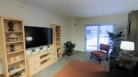 This single-floor space features a welcoming living area characterized by neutral, light-colored walls and soft carpeted flooring that contribute to a calm, understated ambiance. The room is furnished with a light wood media console housing a large flat-screen television, complemented by a matching bookcase unit with open shelving and drawer storage, offering practical organization. A potted leafy plant adds a touch of natural warmth to the corner of the room. A comfortable accent chair in brown upholstery is positioned near a large sliding glass door, which serves as the room's primary source of natural light. The sliding door opens to an exterior space and features a sheer curtain panel that softens the incoming daylight while maintaining an airy feel. A ceiling vent indicates a central climate control system. The overall layout is open and functional, with a straightforward design aesthetic that emphasizes clean lines and a neutral color palette. The space is accented with small potted plants atop the media unit, adding subtle decorative interest throughout the living area.