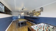 This single-floor game room is designed as a dedicated entertainment and social space, combining functional amenities with distinctive industrial-inspired décor. The room features warm hardwood-style flooring that contrasts with the bold two-tone wall treatment — crisp white upper walls meet deep navy blue wainscoting, creating a sharp, modern aesthetic grounded in classic design sensibility.

At the center of the space sits a full-size pool table illuminated by a pendant light fixture with an angular shade, adding a focused, purposeful lighting element. A foosball table is positioned nearby, offering additional recreational options. Along one wall, a long, dark-topped bar counter is lined with industrial metal stools, providing ample seating for spectators or casual gatherings.

The standout design feature is a sculptural wall installation composed of exposed metal pipes and fittings, arranged in an artistic, geometric pattern above the bar area. Recessed lighting throughout the ceiling ensures even illumination across the room. A windowed door at the far end allows natural light to filter in, enhancing the overall ambiance of this well-appointed, recreation-focused space.