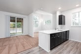 This single-story residence presents a thoughtfully designed open-concept layout where the kitchen and living areas flow seamlessly together. The kitchen is well-appointed with dark espresso cabinetry contrasting against crisp white countertops, stainless steel refrigerator, black appliances including an electric range and built-in microwave, and a stainless steel undermount sink. A peninsula with an integrated sink extends into the living space, offering additional prep surface and casual seating potential. Light wood-style flooring runs through the kitchen, transitioning to plush neutral carpet in the living and sleeping areas.

The living room benefits from a vaulted ceiling and multiple windows that invite natural light, while sliding glass doors open directly onto a covered balcony with pergola-style detailing and outdoor views of surrounding greenery. The bathroom features wood-style flooring, a full tub-shower combination, a toilet, and a dark-cabinetry vanity with a white countertop. A generously sized walk-in closet outfitted with wire shelving systems provides ample storage. The overall palette of neutral tones, recessed lighting, and clean finishes creates a bright, modern ambiance throughout.