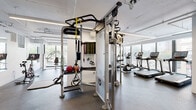 Located at 4557 N Sheridan Rd in Chicago's Uptown neighborhood, this single-floor fitness center offers a comprehensive workout environment designed for a wide range of training styles. The space features a clean, modern aesthetic with white walls, exposed ceiling infrastructure, and durable rubber flooring throughout.

Large floor-to-ceiling windows line multiple walls, flooding the interior with natural light and offering views of the surrounding streetscape and greenery. The cardio zone is well-appointed with multiple Life Fitness treadmills, elliptical machines, and stationary bikes positioned to take full advantage of the outdoor views.

A dedicated strength training area houses a multi-functional cable machine with TRX and resistance band attachments, a full dumbbell rack ranging across multiple weight increments, weight benches, and several selectorized resistance machines for targeted muscle groups. A wall of mirrors enhances the sense of space and supports form monitoring during workouts.

The functional training zone includes stability balls, kettlebells, and medicine balls organized on a dedicated storage rack. A wall-mounted flat-screen display adds an engaging media element to the space. Additional amenities include a water fountain and open floor space ideal for stretching or bodyweight exercises. Linear LED lighting provides consistent, even illumination throughout.