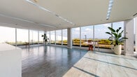 Located at 144 S Harrison St in East Orange, NJ, this thoughtfully designed amenity floor offers a compelling mix of wellness, socializing, and relaxation spaces. Floor-to-ceiling glass walls in the main lounge and corridor flood the interior with abundant natural light while framing sweeping views of a lush rooftop green space and open sky beyond. Large-format tile flooring runs throughout the sun-drenched lounge area, which opens visually to the verdant outdoor terrace, creating a seamless indoor-outdoor ambiance. A sophisticated resident lounge features warm wood flooring, recessed and cove lighting, and clean-lined white seating arranged around a tufted ottoman, conveying a calm, contemporary atmosphere. Adjacent to the lounge is a flexible multi-purpose room finished with patterned carpet tiles and glass partition walls, ideal for private gatherings or meetings. The state-of-the-art fitness center is generously proportioned and divided into distinct zones: a cardio area lined with treadmills and elliptical machines backed by full-length mirrors, and a strength training zone equipped with cable machines, benches, and functional training tools including medicine balls and a rowing machine. Rubber flooring throughout the gym ensures both safety and durability. The entire floor reflects a modern, amenity-rich design tailored for active, community-oriented living.