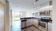 Located at 301 12th Ave in Seattle's vibrant Capitol Hill neighborhood, this one-bedroom, one-bathroom unit offers a thoughtfully designed layout with a clean, contemporary aesthetic throughout. The open kitchen serves as the centerpiece of the living space, featuring a full suite of stainless steel appliances — including a refrigerator, electric range, over-the-range microwave, and dishwasher — paired with dark countertops and sleek white cabinetry that extends to the ceiling for ample storage. Pendant lighting and a track-light fixture illuminate the space stylishly, while a large floor-to-ceiling window floods the area with natural light and provides pleasant outdoor views. Wood-style plank flooring runs throughout the main living and bedroom areas, lending warmth and continuity to the design. The bedroom is a well-proportioned, quiet retreat featuring a sliding closet with natural wood-finish doors and a baseboard heater. A distinctive plywood accent wall adds an organic, modern design touch to the hallway connecting the rooms. The bathroom features terrazzo-style flooring, a vanity with a dark countertop, and a bathtub. The unit's efficient, single-floor layout makes excellent use of its space, offering a functional and modern urban living experience.