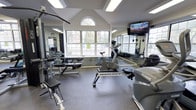 This fitness center is housed in a single-story space distinguished by its vaulted, cathedral-style ceiling that creates an open and airy atmosphere. A prominent arched window at the peak of the gabled wall, flanked by large multi-pane windows on surrounding walls, floods the room with natural light and offers views of the greenery outside. The generous ceiling height enhances the sense of spaciousness throughout the workout area.

The facility is outfitted with a well-rounded selection of exercise equipment. Cardio options include a stationary upright bike and an Octane-brand machine, while strength training is supported by a BodyCraft functional trainer and a dedicated bench press station with a weight rack. Full-length mirrors line the walls, amplifying the sense of space and allowing users to monitor their form during workouts. A wall-mounted flat-screen television provides entertainment during exercise sessions.

The flooring consists of durable rubber gym matting, practical for both equipment placement and user safety. Recessed and panel lighting supplements the natural light, ensuring the space remains well-lit throughout the day. The overall ambiance is clean, functional, and inviting.