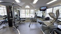This fitness center is housed in a single-story space distinguished by its vaulted, cathedral-style ceiling that creates an open and airy atmosphere. A prominent arched window at the peak of the gabled wall, flanked by large multi-pane windows on surrounding walls, floods the room with natural light and offers views of the greenery outside. The generous ceiling height enhances the sense of spaciousness throughout the workout area.

The facility is outfitted with a well-rounded selection of exercise equipment. Cardio options include a stationary upright bike and an Octane-brand machine, while strength training is supported by a BodyCraft functional trainer and a dedicated bench press station with a weight rack. Full-length mirrors line the walls, amplifying the sense of space and allowing users to monitor their form during workouts. A wall-mounted flat-screen television provides entertainment during exercise sessions.

The flooring consists of durable rubber gym matting, practical for both equipment placement and user safety. Recessed and panel lighting supplements the natural light, ensuring the space remains well-lit throughout the day. The overall ambiance is clean, functional, and inviting.