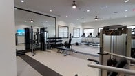 This fitness center is designed to support a comprehensive workout experience within a single-level layout. The space is thoughtfully organized into distinct functional zones, separating cardio and strength training areas for efficient use. Along one side, a row of treadmills equipped with individual monitors provides dedicated cardio space, while a dumbbell rack stocked with a full range of free weights anchors the strength training section. A multi-use cable machine adds versatility for various resistance exercises, and an adjustable workout bench offers additional options for targeted training. Large mirrored panels line one wall, visually expanding the room and allowing users to monitor their form during exercise. Recessed lighting and multiple ceiling fans maintain a bright and comfortable atmosphere throughout. Several windows allow natural light to filter into the space, contributing to an open and airy feel. The flooring features a durable, cushioned surface suited for both weighted and bodyweight movements. The overall design is clean and modern, with neutral tones and a well-organized layout that creates a functional and welcoming environment for fitness enthusiasts of all levels.