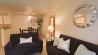 This single-floor residence offers a cozy and well-appointed layout designed for comfortable living. The living area serves as the heart of the space, featuring plush dark upholstered seating arranged around a circular glass coffee table, creating an intimate setting ideal for relaxing or entertaining. Warm ambient lighting from a classic table lamp and a ceiling fan with integrated lighting casts a soft, welcoming glow throughout the room. Decorative accents, including accent pillows, fresh florals, and a stylish sunburst mirror, add personality and charm to the space. Neutral-toned walls and light carpeting contribute to the overall sense of warmth and openness. A tall indoor bamboo plant introduces a natural element, enhancing the inviting atmosphere. Adjacent to the living area, the dining space accommodates a dark wood dining table and chairs, illuminated by the same ceiling fan fixture, making it a seamless extension of the main living area. A doorway visible from the living room hints at additional rooms beyond, suggesting a practical and connected floor plan. Overall, this residence combines functional design with warm, curated décor to create a welcoming and livable environment.