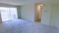 This single-floor residential unit offers a clean, functional layout centered around a spacious living area finished with plush, light-toned wall-to-wall carpeting that adds warmth underfoot. The room is bathed in natural light courtesy of a large sliding glass door fitted with vertical blinds, which provides both privacy control and access to an outdoor area. Neutral, cream-toned walls create a bright and airy atmosphere throughout the space.

A hallway extends from the main living area, connecting to additional rooms and providing a sense of depth to the overall floor plan. Ceiling-mounted lighting in the hallway ensures the transition between spaces remains well-lit and welcoming. A wall-mounted thermostat is conveniently positioned in the living area, offering easy climate control.

The open layout of the living area allows for flexible furniture arrangements, making it easy to personalize the space. The consistent flooring and color tones throughout contribute to a cohesive, understated aesthetic. Overall, the unit presents a straightforward, move-in-ready environment suitable for a variety of living arrangements.