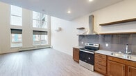 This two-floor residence offers a thoughtfully designed layout with modern finishes and an open, airy atmosphere. The living area is a standout, boasting dramatic double-height ceilings and floor-to-ceiling windows that flood the space with natural light and frame views of the surrounding urban environment. Light wood-tone flooring runs throughout, complementing the clean white walls and creating a bright, cohesive aesthetic. A wall-mounted mini-split system provides efficient climate control alongside baseboard heating.

The kitchen is well-appointed with warm wood cabinetry, open floating shelves, a stainless steel appliance suite including a refrigerator, electric range, dishwasher, and microwave, as well as a stainless steel range hood and a grey tile backsplash that adds texture and visual interest.

The bathroom features matching wood cabinetry, a single vanity with a grey countertop, a large mirror, and wood-look flooring for a clean, functional finish. A dedicated laundry closet houses a stacked washer and dryer unit, offering added convenience. A spacious walk-in closet with wire shelving and hanging rods provides generous storage. The overall ambiance is modern and minimalist, offering a clean canvas well-suited to a variety of living arrangements.