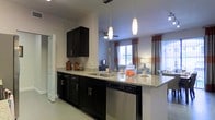 This single-floor residence showcases a thoughtfully designed open-concept layout that seamlessly connects the kitchen and living areas. The kitchen is a standout feature, boasting dark espresso cabinetry with sleek silver hardware that creates a striking contrast against the light granite countertops. A peninsula with an integrated sink and a stainless steel dishwasher provides ample workspace and casual seating, while two elegant pendant lights hang overhead, adding a warm, focused glow to the space. Light tile flooring runs throughout, contributing to a clean, cohesive aesthetic. The living area beyond the kitchen benefits from generous natural light streaming through multiple windows dressed with bold, color-blocked curtains in orange and gray, lending a vibrant touch to the space. A ceiling fan keeps the area comfortable, and the open sightlines between the kitchen and living room make the overall layout feel expansive and inviting. The neutral wall palette and well-coordinated design elements throughout give this residence a modern, move-in-ready appeal that balances both style and everyday functionality.