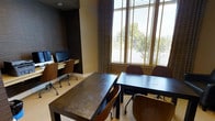 This single-floor business center offers a thoughtfully designed environment tailored for work and collaboration. The space is anchored by a built-in computer workstation along one wall, equipped with desktop monitors and ergonomic rolling chairs, making it ideal for individual tasks requiring focused attention. Complementing this setup are two dark-toned rectangular tables with matching wooden chairs, providing ample space for group meetings, study sessions, or independent work. The room's design strikes a balance between professional and inviting, featuring textured dark wall panels that add depth and sophistication to the interior. Large windows fitted with horizontal blinds and accented by elegant floor-length drapes allow generous natural light to filter in while maintaining privacy, and offer views of lush greenery outside. The neutral-toned flooring and warm wall colors create a cohesive and comfortable atmosphere. The overall layout is practical yet polished, making this space well-suited for a range of professional and collaborative activities.