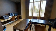 This single-floor business center offers a thoughtfully designed environment tailored for work and collaboration. The space is anchored by a built-in computer workstation along one wall, equipped with desktop monitors and ergonomic rolling chairs, making it ideal for individual tasks requiring focused attention. Complementing this setup are two dark-toned rectangular tables with matching wooden chairs, providing ample space for group meetings, study sessions, or independent work. The room's design strikes a balance between professional and inviting, featuring textured dark wall panels that add depth and sophistication to the interior. Large windows fitted with horizontal blinds and accented by elegant floor-length drapes allow generous natural light to filter in while maintaining privacy, and offer views of lush greenery outside. The neutral-toned flooring and warm wall colors create a cohesive and comfortable atmosphere. The overall layout is practical yet polished, making this space well-suited for a range of professional and collaborative activities.