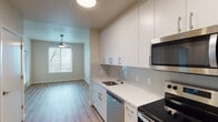 Located at 3158 SW 12th Ave in Portland, Oregon, this single-floor unit offers a clean, modern aesthetic with thoughtfully designed living spaces. The kitchen is a standout feature, equipped with flat-front white cabinetry extending floor to ceiling, white quartz countertops, a glass tile backsplash, and a full suite of stainless steel appliances including an electric range, built-in microwave, dishwasher, and refrigerator. The kitchen flows seamlessly into the open living area, which is finished with warm wood-tone luxury vinyl plank flooring and features a ceiling fan for added comfort. A wall-mounted mini-split system provides efficient climate control. Natural light enters through large windows fitted with horizontal blinds, offering views of surrounding greenery. The bedroom is a well-proportioned, quiet retreat with soft carpeting, a ceiling fan, and its own window with outdoor views. A hallway connects the main living areas to the bathroom, which includes a single-sink vanity with white cabinetry, a large mirror, and a full tub-shower combination with tile surround. The overall design is cohesive and contemporary, with neutral tones, recessed lighting, and durable finishes suited for everyday living.