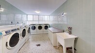 This well-appointed communal laundry facility spans a generously sized single floor, thoughtfully designed to accommodate a high volume of use with efficiency and comfort in mind. The space is divided into two interconnected rooms, both finished with classic sage green ceramic wall tiles that lend a clean, institutional character throughout.

The main room is anchored by a central granite-topped folding island, providing ample surface area for sorting and folding laundry. Rows of modern front-load Maytag washers and dryers line the perimeter walls, offering substantial capacity for residents. Informational signage is posted along the walls, keeping users informed on machine operation and facility guidelines.

A secondary room provides additional machines, including a standalone washer, as well as a freestanding utility sink with a countertop work surface — ideal for hand-washing or pre-treating garments. An electrical panel is also accessible within this area.

Natural light filters in through glass block windows positioned along the upper walls, brightening the space without compromising privacy. Overhead flush-mount light fixtures provide consistent illumination throughout. The durable terrazzo flooring, speckled in neutral tones, ties the rooms together and is well-suited for the demands of a high-traffic laundry environment. The overall layout is practical, open, and easy to navigate.
