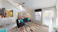 This single-floor residence offers a welcoming and well-designed living environment highlighted by striking vaulted ceilings that lend an expansive, airy feel to the main living area. A sleek ceiling fan is centrally mounted, providing both function and style beneath the textured ceiling. The living room is generously sized, featuring plush wall-to-wall carpeting that adds warmth underfoot. Natural light flows in through multiple windows fitted with blinds, creating a bright and comfortable atmosphere throughout the day. A glass-paneled door provides additional light and easy access to the outdoors. The open layout connects the living room seamlessly to the dining area, which is visible in the background and offers a cheerful, inviting space for meals and gatherings. A pass-through or partial wall hints at a kitchen beyond, reinforcing the home's open and connected floor plan. Neutral-toned walls serve as a versatile backdrop throughout the space, complementing the light-filled rooms. The combination of vaulted ceilings, natural light, and a flowing layout makes this residence feel spacious and comfortable, well-suited for both everyday living and entertaining.