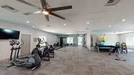This fitness center is a comprehensive, single-floor facility designed to support a wide range of workout routines. The space is anchored by a bold, typographic mural spanning an entire wall, featuring motivational fitness-related words in teal, green, and dark tones, adding an energetic and inspiring atmosphere throughout.

The main workout area is generously proportioned, featuring patterned carpet tiles with green accent stripes that define the open floor plan. Cardio equipment includes multiple treadmills, elliptical machines, and stationary bikes, with the treadmill section accompanied by a wall-mounted flat-screen television and flanked by windows that allow natural light to fill the space. A ceiling fan provides additional air circulation over the cardio zone.

The strength training section offers a cable/functional trainer machine, a weight bench, and a well-stocked free weights area featuring a full rack of dumbbells and a set of kettlebells in varying sizes. A floor-to-ceiling mirror wall expands the sense of space and assists with form during workouts.

Additional equipment includes a medicine ball rack with an assortment of colorful balls and resistance accessories. Windows throughout the facility provide views of the surrounding outdoor area, including a pool and lounge space visible from within. Recessed lighting and neutral wall tones contribute to a clean, modern aesthetic throughout the facility.