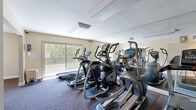 Located in Van Nuys, Los Angeles, this fitness center offers a well-organized and functional workout space across a single floor. The exercise room is equipped with multiple elliptical trainers, treadmills, and a multi-station strength machine, providing a comprehensive range of cardio and resistance training options. Wall-to-wall mirrors line key sections of the room, visually expanding the space and allowing users to monitor their form during workouts. The flooring throughout the exercise area is fitted with durable, cushioned carpet, offering comfort underfoot during extended training sessions. Natural light floods the room through large sliding glass doors that face a lush, green outdoor area, creating a pleasant and energizing atmosphere. An additional window further enhances the bright, airy feel of the space. Overhead fluorescent lighting ensures consistent illumination throughout the day. A short hallway connects the exercise room to the entry, providing a clean transition into the fitness area. The neutral, warm-toned walls and simple, uncluttered layout contribute to a focused and practical environment well-suited for daily exercise routines.