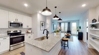 This thoughtfully designed single-floor residence showcases a contemporary open-concept layout that seamlessly connects the kitchen, dining, and living areas. The kitchen serves as a central focal point, featuring a large granite-topped island with an undermount sink and matte black faucet, white shaker-style cabinetry, classic subway tile backsplash, and stainless steel appliances including a side-by-side refrigerator. Three pendant lights with industrial-style black shades illuminate the island, adding a stylish contrast to the bright, white cabinetry. Wood-look flooring runs throughout the main living areas, lending warmth and continuity to the space.

The living room is bright and inviting, benefiting from multiple windows and a glass door that opens to a private balcony, allowing generous natural light to fill the room. The bedrooms are carpeted for comfort, each offering multiple windows with outdoor views and ceiling fans. Walk-in closets with wire shelving systems provide practical storage solutions. The bathroom features clean white subway tile surrounding the tub and shower, paired with a granite-topped vanity and wood-look flooring, creating a crisp, modern aesthetic throughout.