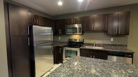 This single-floor space is designed with a contemporary aesthetic that emphasizes functionality and style. The kitchen serves as a standout feature, showcasing rich, dark espresso-toned cabinetry that extends to the ceiling, maximizing storage capacity. The cabinetry is complemented by striking granite countertops with a bold, varied pattern in deep blues and grays, lending a sophisticated character to the space. A matching granite kitchen island or peninsula provides additional workspace and reinforces the cohesive design language throughout.

The kitchen is fully equipped with a complete suite of stainless steel appliances, including a full-size refrigerator, an electric range with oven, an over-the-range microwave, and a stainless steel sink. The appliances are seamlessly integrated into the cabinetry layout, creating a streamlined and polished look. Recessed lighting in the ceiling provides bright, even illumination across the workspace.

The neutral gray walls serve as a clean backdrop that ties the dark cabinetry and metallic finishes together harmoniously. The light-toned flooring contrasts effectively with the darker tones of the cabinetry, adding balance and visual interest. Overall, the space presents a modern, well-coordinated interior that is both practical and visually refined.
