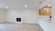 This single-floor space offers a clean, understated design centered around a welcoming living area. The room is finished with soft, neutral-toned carpeting that extends throughout, contributing to a warm and comfortable atmosphere. A built-in fireplace with a sleek black surround serves as a natural focal point along one wall, adding both character and function to the space. The crisp white walls and recessed lighting keep the interior feeling bright and airy. Adjacent to the living area, a built-in counter pass-through connects to a kitchen space, complemented by warm-toned wooden upper cabinetry. This layout creates a sense of openness while maintaining a natural flow between the living and kitchen areas. The visible HVAC vent in the ceiling suggests a central climate control system, adding to the practical comfort of the space. Overall, the design is minimalist and move-in ready, with thoughtful architectural details that balance simplicity with functionality, making it a versatile and inviting environment.