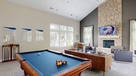 Located at 4901 Warner Rd in Columbus, Ohio, this single-story community clubhouse offers a well-appointed gathering space designed for both recreation and relaxation. The main game room is the centerpiece of the facility, boasting soaring vaulted ceilings that lend an airy, open feel to the space. A full-size billiard table anchors the room, complemented by plush carpeting, barn-style sliding doors, and abundant natural light streaming through large windows that frame outdoor views.

Adjacent to the game room is an elegantly furnished lounge area featuring a dramatic floor-to-ceiling stacked stone fireplace with a mounted flat-screen television above the mantle. Tufted leather seating, a glass-top coffee table, and curated artwork create a sophisticated yet comfortable atmosphere. Large windows continue to bring in natural light, with views of the surrounding landscape and pool area.

A quieter sitting room off the main space offers a more intimate retreat, furnished with a neutral-toned sofa and armchair, decorative greenery, and tasteful wall art. The facility also includes a functional galley-style kitchen equipped with granite countertops, wood cabinetry, stainless steel sink, and black appliances, making it ideal for hosting community events and gatherings.