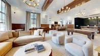 Located at 2604 Secretariat Blvd in Grove City, Ohio, this single-floor amenity space offers a generously sized, thoughtfully designed clubroom experience. The interior showcases a refined aesthetic, combining warm walnut wood wainscoting with crisp white walls, gleaming large-format tile flooring, and elegantly styled brass and gold chandeliers that illuminate the space throughout.

The main gathering area features an open-plan layout with multiple distinct zones. A plush lounge area is anchored by rich amber and neutral upholstered sofas and armchairs, complemented by a large mural depicting a lush golf course landscape. Floor-to-ceiling windows flood this zone with natural light while providing views of an outdoor pool area.

A dedicated game and entertainment zone houses a full-sized billiards table set beneath coffered ceilings with exposed dark wood beams. Adjacent to it, a full golf simulator bay — complete with a projected green turf hitting surface and large display screen — provides an immersive experience. A bar counter with high-top seating and wood-paneled cabinetry serves as a natural social hub. Framed artwork and carefully curated décor add to the polished, club-like atmosphere throughout this versatile and well-appointed amenity space.