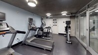 This compact yet fully outfitted fitness center is designed to support a comprehensive workout routine within a single, efficiently arranged space. The room features an exposed concrete ceiling paired with clean white and soft blue-grey walls, giving the space a modern, utilitarian aesthetic. Durable rubberized flooring throughout provides traction and cushioning for a wide range of exercises.

The cardio section is anchored by Matrix-brand equipment, including a treadmill, an elliptical trainer, and an upright stationary bike, each equipped with an individual display screen for tracking workout metrics. Complementing the cardio offerings is a well-stocked free weights area, featuring a three-tier dumbbell rack with a full range of weights and a colorful collection of stability balls in multiple sizes. A tiered kettlebell rack adds further versatility for functional training.

A wall-mounted exercise mat provides a designated space for stretching, core work, or bodyweight exercises. Flush-mounted ceiling lights illuminate the room evenly, and large glass panels along one wall allow natural light to filter in from an adjacent area, enhancing the overall brightness of the space. The fitness center offers a focused and functional environment suitable for a variety of training needs.