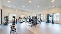 The Statesman's fitness center is a thoughtfully designed, single-floor facility that combines functionality with refined aesthetics. The entrance corridor features light hardwood flooring, a whitewashed brick accent wall adorned with an illuminated cursive logo sign, and seating, setting an inviting tone before entering the workout space.

The main cardio and strength area is expansive and bright, with high ceilings, recessed lighting, and decorative pendant fixtures. Large steel-framed windows along multiple walls invite abundant natural light and outdoor views. This room houses a comprehensive selection of Matrix-branded strength machines, cable systems, treadmills, ellipticals, and stair climbers arranged across durable flooring.

An adjoining free weights room features blue-painted walls, adjustable benches, a dumbbell rack, stationary bike, medicine balls, resistance bands, and stability equipment. A separate studio area provides an open, airy environment with soaring ceilings, direct exterior access, and dual mounted screens for on-demand workout programming. Rowing machines are also available in this space.

A full glass wall overlooking a dramatic double-height lobby with woven pendant lights and floor-to-ceiling windows adds a striking architectural element to the overall fitness center experience.