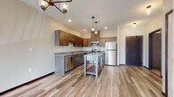 This well-appointed single-floor unit offers a thoughtfully designed open-concept layout that seamlessly blends the kitchen and living areas. The kitchen is a standout feature, showcasing rich dark wood cabinetry, granite countertops, and a full suite of stainless steel appliances including a refrigerator, range, microwave, and dishwasher. A freestanding kitchen island provides additional workspace and storage. Stylish pendant lights hang above the kitchen area, complementing the sputnik-style chandelier that adds a modern touch to the adjacent living space. Light-toned hardwood flooring extends throughout the main living areas, creating a warm and cohesive aesthetic. The bedroom features plush carpeting, a ceiling fan, a window with outdoor views, and baseboard heating, offering a comfortable and functional retreat. Additional rooms, also carpeted, provide versatile living space. The bathroom is finished with the same quality materials found throughout the unit, including hardwood-style flooring, a granite-topped vanity with matching dark wood cabinetry, and a walk-in shower with grab bar accessibility features. Dark-framed doors and trim add a refined contrast to the neutral, light-toned walls throughout the space.