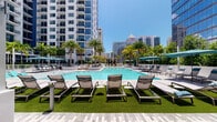 This expansive outdoor amenity deck offers a resort-inspired retreat within an urban high-rise setting. At its center sits a generously sized swimming pool with crystal-clear blue water, surrounded by a sophisticated hardscape of light-toned stone pavers that lend an elegant, contemporary feel to the space. Rows of modern mesh lounge chairs are arranged poolside on vibrant artificial turf sections, providing comfortable spots to relax and soak up the abundant natural sunlight. Teal market umbrellas are strategically positioned throughout the deck to offer shade and a pop of color against the bright surroundings. Lush tropical landscaping, including tall palm trees and dense, verdant plantings in crisp white raised planters, frames the space and adds a natural, resort-like ambiance. Private cabana-style seating areas are nestled among the greenery, each enclosed by white pergola structures and furnished with wicker lounge chairs and small side tables — offering an intimate retreat from the open pool area. The entire deck is set against a dramatic backdrop of gleaming glass high-rise towers, reinforcing its urban luxury character. The combination of thoughtful landscaping, varied seating options, and open sky exposure makes this pool deck an exceptional outdoor living destination.