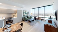 This contemporary single-floor residence is defined by its striking floor-to-ceiling windows that frame panoramic city views, flooding the open-plan living and dining area with abundant natural light. The living space features light hardwood flooring throughout, clean white walls, and a seamless connection between the lounge and kitchen areas, creating an airy, cohesive environment.

The kitchen is outfitted with modern flat-front cabinetry in a soft grey finish, stainless steel appliances including a large French door refrigerator and a gas range, white countertops, and recessed lighting overhead. A live-edge wood dining table adds a warm, organic contrast to the otherwise sleek aesthetic.

The bedroom is a tranquil retreat, benefiting from its own floor-to-ceiling window with direct city views. The room is simply appointed with neutral tones, quality furnishings, and curated artwork, and connects directly to the full bathroom.

The bathroom is finished with hexagonal tile flooring, large-format stone-look wall tiles in the soaking tub and shower surround, a modern floating vanity with an illuminated mirror, and crisp white countertops — offering a spa-like atmosphere.

Throughout, the space maintains a cohesive modern design language marked by clean lines, quality materials, and a neutral palette accented by thoughtful pops of color.