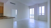 This single-floor space showcases a clean, modern design characterized by an all-white interior palette that enhances its bright and airy atmosphere. The open-concept living area is finished with large-format white tile flooring laid in a staggered pattern, contributing to a seamless and expansive feel throughout the space. Elevated ceilings add a sense of volume to the main living area, while crisp white walls provide a neutral canvas throughout.

A standout feature is the floor-to-ceiling sliding glass door that opens directly onto a balcony, offering open outdoor views and flooding the interior with abundant natural light. An additional window positioned to the right further amplifies the brightness of the space. The balcony itself is enclosed by a white railing, suggesting elevated positioning with scenic surroundings.

Adjacent to the living area, a partial view of a kitchen counter with a stone or granite surface hints at a functional and stylish culinary space. A doorway visible from the main living area provides access to additional rooms. The HVAC vent integrated into the upper wall indicates a central climate control system. Overall, the space presents a light-filled, contemporary environment with practical layout and quality finishes.