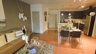This single-floor residence offers a thoughtfully designed open-concept layout that connects the living, dining, and kitchen areas in one cohesive flow. Warm hardwood flooring extends throughout the main living space, lending a sense of continuity and natural warmth to the interior.

The living area is anchored by a plush sofa and a glass coffee table atop a patterned area rug, complemented by decorative accents including a sculptural white stag head mounted on a rich, dark feature wall. The contrast between the earthy brown accent wall and the crisp white walls throughout creates a modern, layered aesthetic.

The dining area sits adjacent to the living room and features a sleek rectangular table paired with contemporary woven leather chairs. Pendant lighting drops from above, casting focused, ambient light over the dining space and enhancing its modern character.

The kitchen is finished with dark cabinetry, stainless steel appliances including a refrigerator and microwave, and a clean, efficient layout. Pendant lights above the counter add a polished touch while maintaining visual harmony with the rest of the space.

Two interior doors lead to additional rooms, maintaining a sense of privacy without interrupting the open feel of the main living area. Overall, the space presents a clean, modern, and well-appointed interior suitable for comfortable everyday living.