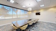 Located at 2701 Neabsco Common Pl in Woodbridge, VA, this professional conference room offers a polished and functional environment designed for productive meetings. The space centers around a large, sleek conference table surrounded by comfortable upholstered swivel chairs, providing ample seating for a full team. A dropped tile ceiling with recessed lighting and a decorative pendant fixture creates a bright, well-lit atmosphere, while a wall-mounted flat-screen display adds modern utility for presentations. One wall features large windows fitted with roller shades, allowing natural light to filter in while offering views of the surrounding outdoor greenery. A built-in credenza with storage cabinets runs along one wall, topped with decorative accents including a teal vase and vibrant abstract artwork that adds a touch of color and character to the otherwise neutral, cream-toned interior. Glass-paneled double doors provide an elegant entry point and visual connection to the adjacent corridor. The patterned carpet in muted blue and gray tones anchors the room with a contemporary aesthetic. This conference room balances professional functionality with thoughtful design details, making it a versatile and inviting space for meetings, presentations, and collaborative work sessions.