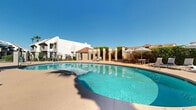 Located at 400 N 96th Ave in Tolleson, Arizona, this outdoor amenity space showcases a generously sized freeform swimming pool as its centerpiece. The pool's vivid turquoise water is complemented by a wide, open concrete surround that provides ample room for lounging and recreation under the Arizona sun. Surrounding the pool deck is a thoughtfully arranged collection of lounge chairs and patio seating grouped beneath striped umbrellas, offering shaded areas for relaxation and socializing. Architectural pergola-style columns line portions of the deck, adding a structured, resort-inspired aesthetic to the outdoor environment. Lush green hedges and mature palm trees frame the perimeter, providing both privacy and a vibrant contrast to the neutral stucco exteriors of the adjacent multi-story buildings, which feature warm terra cotta roof tiles characteristic of Southwest regional architecture. A gated entry with black iron railings leads into the space, ensuring a secure and well-defined boundary. The overall ambiance is that of a well-maintained, community-oriented outdoor retreat, designed to maximize comfort and enjoyment in the warm desert climate of the greater Phoenix area.