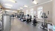 Located at 5605 Holly Grove Way in Knoxville, Tennessee, this fitness center offers a comprehensive workout environment housed in a single, open-plan floor. The space is characterized by warm beige walls accented with crisp white crown molding, rubber-floored workout areas, and a row of arched windows fitted with plantation shutters that allow generous natural light to fill the room.

The cardio section is well-stocked with multiple treadmills, stationary bikes, an air bike, and elliptical machines, all arranged to maximize floor space and ease of movement. Strength-training options include cable machines, a multi-station weight system, a rowing machine, and a dedicated flat bench, providing a thorough range of resistance exercises. A full-length wall mirror on one end of the room visually expands the space and allows users to monitor their form during workouts. A wall-mounted flat-screen TV adds an entertainment element to the cardio area.

Practical amenities include a small beverage refrigerator, a water station, and posted fitness center rules near the entrance. Bright overhead fluorescent lighting supplements the natural light from the windows. The arched entryway door with an illuminated exit sign contributes to the room's classic architectural character. Overall, the space presents a clean, functional, and inviting environment well-suited for a well-rounded fitness routine.