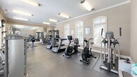 Located at 5605 Holly Grove Way in Knoxville, Tennessee, this fitness center offers a comprehensive workout environment housed in a single, open-plan floor. The space is characterized by warm beige walls accented with crisp white crown molding, rubber-floored workout areas, and a row of arched windows fitted with plantation shutters that allow generous natural light to fill the room.

The cardio section is well-stocked with multiple treadmills, stationary bikes, an air bike, and elliptical machines, all arranged to maximize floor space and ease of movement. Strength-training options include cable machines, a multi-station weight system, a rowing machine, and a dedicated flat bench, providing a thorough range of resistance exercises. A full-length wall mirror on one end of the room visually expands the space and allows users to monitor their form during workouts. A wall-mounted flat-screen TV adds an entertainment element to the cardio area.

Practical amenities include a small beverage refrigerator, a water station, and posted fitness center rules near the entrance. Bright overhead fluorescent lighting supplements the natural light from the windows. The arched entryway door with an illuminated exit sign contributes to the room's classic architectural character. Overall, the space presents a clean, functional, and inviting environment well-suited for a well-rounded fitness routine.