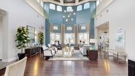Located at 6975 Sierra Club Circle in Naples, Florida, this single-story community clubhouse delivers a sophisticated and welcoming atmosphere through thoughtful architectural design and upscale finishes. The centerpiece of the space is a dramatic grand lounge featuring soaring double-height ceilings, bold teal accent walls, and an abundance of natural light streaming through floor-to-ceiling windows and transom windows positioned at the upper clerestory level. A striking sputnik chandelier anchors the room, complementing the rich hardwood flooring that flows throughout the entire space. Expansive views of the surrounding community pool and grounds are visible through the oversized windows, further enhancing the open, airy ambiance. The lounge is furnished with refined seating arrangements, including velvet wingback chairs, plush sofas, and gold-accented glass coffee tables, creating an inviting area for socializing or relaxing. A dedicated business center offers multiple workstations along a built-in desk, accompanied by comfortable seating and warm pendant lighting — ideal for residents working remotely. The fully equipped kitchen features sleek sage-toned cabinetry, white quartz countertops, and stainless steel appliances. French doors provide seamless access to the outdoor areas, completing this polished and functional community hub.