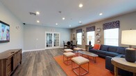 This single-story communal lounge offers a thoughtfully designed open-plan layout that balances comfort and functionality. The space features hardwood-style flooring throughout, complemented by neutral gray walls that provide a clean, modern backdrop. Recessed lighting illuminates the room evenly, creating a bright and welcoming atmosphere.

The lounge is divided into distinct functional zones. A seating area anchored by a navy blue sofa with orange accent pillows sits atop a bold orange area rug, accompanied by a set of upholstered ottomans with metallic frames and a hairpin-leg coffee table. This arrangement encourages casual conversation and relaxation. Decorative silver platters and patterned valances above the windows add a curated, stylish touch.

A dining area toward the rear of the room features a round table with dark chairs, suitable for small group gatherings or meetings. French doors with glass panes connect to an adjacent room, offering visual openness while maintaining separation between spaces. Natural light filters in through multiple windows, enhancing the room's airy feel. The overall ambiance is contemporary and inviting, well-suited for a shared community environment.