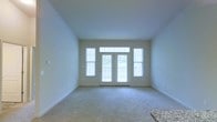 This single-floor space offers a clean, modern layout designed to maximize comfort and natural light. The living area is a standout feature, characterized by soft, wall-to-wall carpeting and crisp white walls that create a bright, airy atmosphere. A set of French doors serves as the focal point of the main living space, flanked by large windows on either side that flood the room with abundant natural light. The doors open to a private balcony, providing a seamless connection between the interior and the outdoors. A transom window above the French doors further enhances the sense of openness and light. The neutral color palette throughout — from the light-toned walls to the understated carpeting — lends the space a versatile, move-in-ready appeal. Granite countertop detailing, partially visible at the edge of the frame, hints at quality finishes in the adjoining areas. The overall ambiance is calm, clean, and welcoming, making the space well-suited for a variety of uses and furnishing styles.