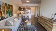 This single-floor residence showcases an open-concept layout that connects the living area and kitchen into one cohesive, functional space. Dark hardwood flooring runs throughout, lending a warm and polished aesthetic that ties the rooms together seamlessly.

The living area is comfortably sized, anchored by a light gray sofa accented with patterned throw pillows in shades of blue and orange. A navy geometric-patterned area rug defines the seating zone, while a glass-and-metal coffee table serves as a stylish centerpiece. A rustic wood credenza along the far wall adds textural contrast to the contemporary surroundings.

The kitchen features dark espresso cabinetry paired with light countertops, pendant lighting above the breakfast bar, and stainless steel appliances including a full-size refrigerator. Bar-height stools at the counter provide casual dining seating.

A tray ceiling in the living area adds a subtle architectural detail overhead. Tucked neatly into an alcove along one wall is a stacked washer and dryer unit, offering the convenience of in-unit laundry. The overall ambiance is modern and well-appointed, balancing functional design with stylish, contemporary décor.