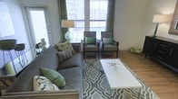 This single-floor space presents a thoughtfully designed living area that balances comfort and contemporary style. The living room serves as the heart of the space, showcasing large windows dressed with blinds and elegant green curtains that flood the room with natural light and offer views of the surrounding urban environment. The light hardwood flooring provides a warm foundation throughout, complementing the room's refined aesthetic. A plush gray sofa anchors the seating arrangement, accented with a mix of patterned and solid green throw pillows that tie the color scheme together. Two classic upholstered armchairs with dark wood frames and green accent pillows face the sofa, creating an inviting conversation area centered around a clean white coffee table with a decorative gold starburst accent. A geometric patterned area rug in shades of green and gray defines the seating zone and adds texture to the space. A stylish green glass table lamp provides ambient lighting, while access to an adjacent area is visible through a glass-paneled door. The overall ambiance is fresh, cohesive, and welcoming, with a carefully curated palette of greens and grays lending the space a polished and modern character.