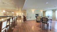 This single-floor space is designed with an open, flowing layout that seamlessly connects multiple functional areas. Rich hardwood flooring extends throughout, anchoring the space with warmth and continuity. A tray ceiling with recessed lighting adds architectural depth overhead, while wall-mounted sconces and standing floor lamps contribute to a layered, ambient lighting scheme.

At the heart of the space sits a stone-framed fireplace, serving as a natural focal point. Elegant upholstered seating is arranged nearby, creating an inviting lounge area suited for conversation or relaxation. Decorative sculptures and tall floral arrangements add tasteful accents throughout.

To one side, a bar-height dining or counter area is furnished with tall chairs, suggesting a casual gathering or dining function. Further back, a console table with table lamps and additional seating define a secondary lounge zone, offering a sense of separation without sacrificing the open feel.

The neutral, earthy wall tones complement the warm flooring and soft furnishings, resulting in a cohesive and refined ambiance throughout the entire space.