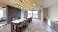 This single-floor space is thoughtfully designed to accommodate both social gatherings and functional activities. The main open area serves as a communal lounge and dining space, anchored by multiple high-top tables with upholstered bar-height chairs arranged beneath elegant gold-framed pendant light fixtures. A striking floor-to-ceiling stacked stone accent wall provides a dramatic backdrop for a sleek built-in fireplace, adding warmth and visual interest to the space. Large windows along one wall allow natural light to fill the interior while providing views of the surrounding streetscape.

Adjacent to the main lounge is a kitchenette equipped with rich dark wood cabinetry, light stone countertops, a stainless steel double sink, dishwasher, full-size refrigerator, and a coffee station — ideal for catering to events or daily use. The flooring throughout is a cohesive wood-look plank in a warm grey tone, lending continuity across all areas.

A separate glass-walled multipurpose room, enclosed by full-height frameless glass panels with black trim, features floor-to-ceiling mirrors and ample natural light, making it well-suited for fitness, dance, or flexible programming needs.