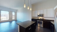 This contemporary single-floor residence showcases a thoughtfully designed open-concept layout that seamlessly connects the kitchen and living areas. Dark hardwood flooring runs throughout the main living spaces, providing a rich contrast against the crisp white walls and soaring ceilings that amplify the sense of space.

The kitchen is a standout feature, equipped with a full suite of stainless steel appliances including a range, over-the-range microwave, dishwasher, and refrigerator. Dark lower cabinetry is paired with white upper cabinets, anchored by dark granite countertops that extend across a central island with pendant lighting above. The island offers additional storage and serves as a natural gathering point.

The bedroom areas are generously proportioned with the same dark hardwood flooring and neutral toned walls. Floor-to-ceiling windows and sliding glass doors flood the rooms with natural light while offering elevated city views of the surrounding urban skyline.

The bathroom maintains the space's modern aesthetic with dark wood vanity cabinetry, a light countertop, a framed mirror with vanity lighting, and large-format grey tile flooring. Clean chrome fixtures and white tile complement the overall design.

Overall, the unit presents a cohesive contemporary style defined by a bold contrast of dark and light elements, abundant natural light, and city-facing outlooks.