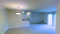 This single-floor residence offers a thoughtfully arranged open-concept layout that connects the main living and kitchen areas seamlessly. The living space is finished with soft, wall-to-wall carpeting in a neutral tone, creating a comfortable and inviting atmosphere. Crisp white walls throughout lend the interior a clean, airy feel, while flush-mounted ceiling lighting ensures consistent illumination across the space.

A defining feature of the layout is the kitchen peninsula, topped with a dark countertop that provides a natural visual boundary between the cooking and living zones. A stylish pendant light hangs above the peninsula, adding a decorative focal point to the open area. The kitchen area is finished with hard flooring, offering a practical contrast to the carpeted living space.

Beyond the peninsula, a hallway leads to additional rooms, with a paneled door visible at the end, suggesting access to private areas of the residence. A wall-mounted HVAC vent is positioned along the baseboard, indicating a built-in climate control system. The overall design is understated and functional, with a neutral palette that provides a versatile backdrop well-suited to a variety of interior styles and furnishing choices.