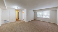 This single-floor residence offers a practical and comfortable layout spread across several well-defined living areas. The kitchen is efficiently arranged in a galley-style configuration, featuring warm honey-toned wood cabinetry, dark granite-look countertops, a stainless steel sink, and a white electric range with overhead ventilation. Light tile flooring and a window fitted with horizontal blinds bring natural light into the space, creating a bright and functional cooking environment. The living area is generously proportioned, finished with wall-to-wall carpeting in a neutral tone that adds warmth underfoot. Large windows dressed with blinds allow ample natural light to filter in, while clean white walls keep the space feeling open and airy. A hallway connects the main living space to the bedroom and bathroom, with a refrigerator visible in the corridor, suggesting convenient storage placement. The bedroom is a comfortable, carpet-floored room with a window providing natural light and direct access to adjacent spaces. The bathroom is clean and straightforward, featuring a full-sized soaking tub with an overhead shower, white subway tile surround, and chrome fixtures. Overall, the unit presents a cohesive, no-frills design that is practical, move-in ready, and well-suited for everyday living.